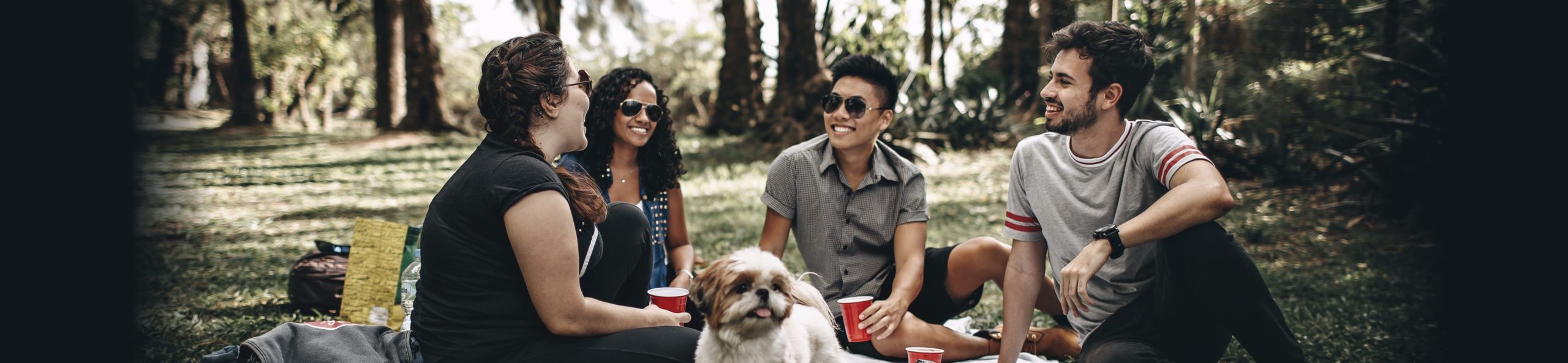 Four people and a dog having a good time at a picnic in a park.