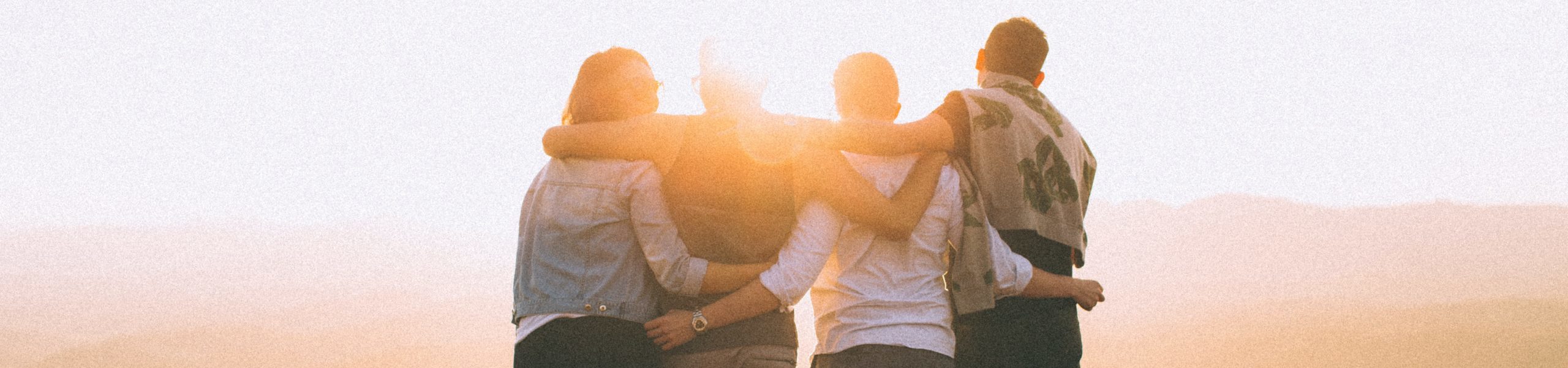 Four people with their backs facing the camera and their arms around each other at sunset.